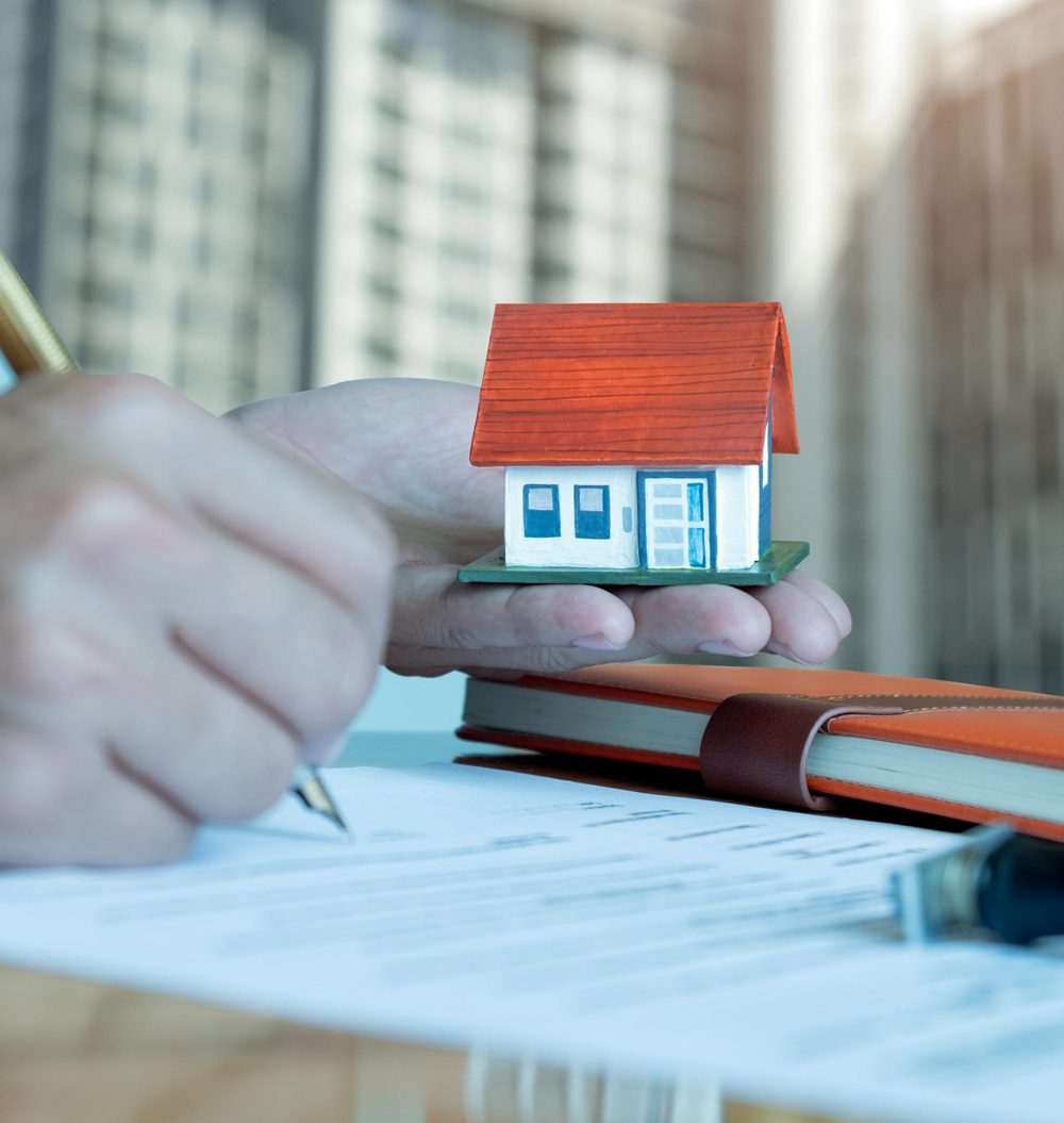 Men have a model house on hand,other hand is using a pen signing contract,background is a blurred condominium image.